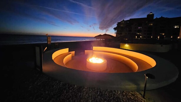 A circular outdoor seating area with a central fire feature, surrounded by a low stone wall, overlooking a body of water and a building with multiple balconies.