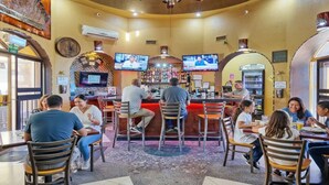 A bar with multiple screens displaying sports events, a counter with a bartender, and several patrons seated at tables.