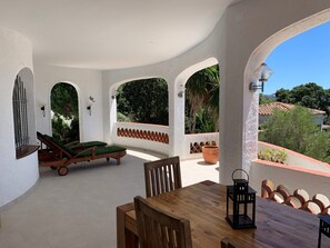 A covered patio with a wooden table and chairs, lounge chairs, and potted plants. - Holiday house in a quiet location, sea view, private pool, near the beach (Roses)