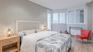 Un dormitorio con cama, mesitas de noche, una silla roja y ventanas grandes.