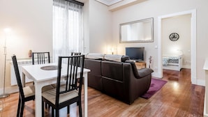 Un salon moderne avec une table à manger, des chaises noires, un canapé marron, un téléviseur à écran plat et un petit lit placé dans un coin.