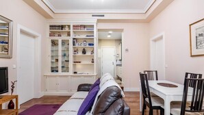 Un salon comprenant un canapé, une table à manger avec des chaises, une bibliothèque intégrée et un téléviseur placé à gauche.