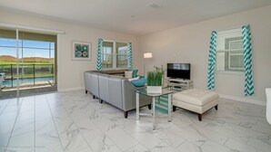 A spacious living room with a grey sofa, a glass coffee table, and a flat-screen TV. There are large windows with chevron-patterned blinds, and a view of a pool and hills in the background. - You and Your Family Will Love This Luxury Mansion on Champions Gate Resort, Orlando Mansion 3726 (Davenport)