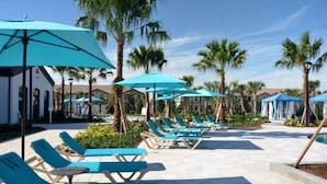 Un espace aménagé au bord de la piscine, avec des chaises longues bleues et des parasols, entouré de palmiers et de bâtiments.