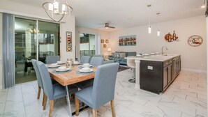 Eine moderne Küche mit einer zentralen Kochinsel, ein Essbereich mit Holztisch und blauen Stühlen sowie ein Wohnbereich mit Sofa und Fernseher.