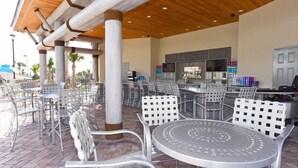 Un patio al aire libre con mesas y sillas redondas, una zona de bar con una televisión y una piscina al fondo.