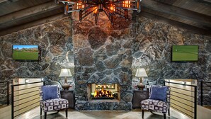 Une cheminée en pierre avec un feu de bois, deux fauteuils à motifs et un téléviseur fixé au mur.