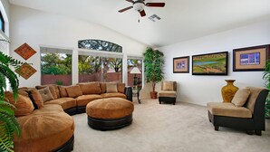Un salon spacieux comprenant un canapé d’angle, un pouf rond, une petite table avec une lampe et un fauteuil. Les murs sont ornés de tableaux encadrés, le plafond est équipé d’un ventilateur et de grandes fenêtres offrent une vue sur les arbres et un mur de briques à l’extérieur.