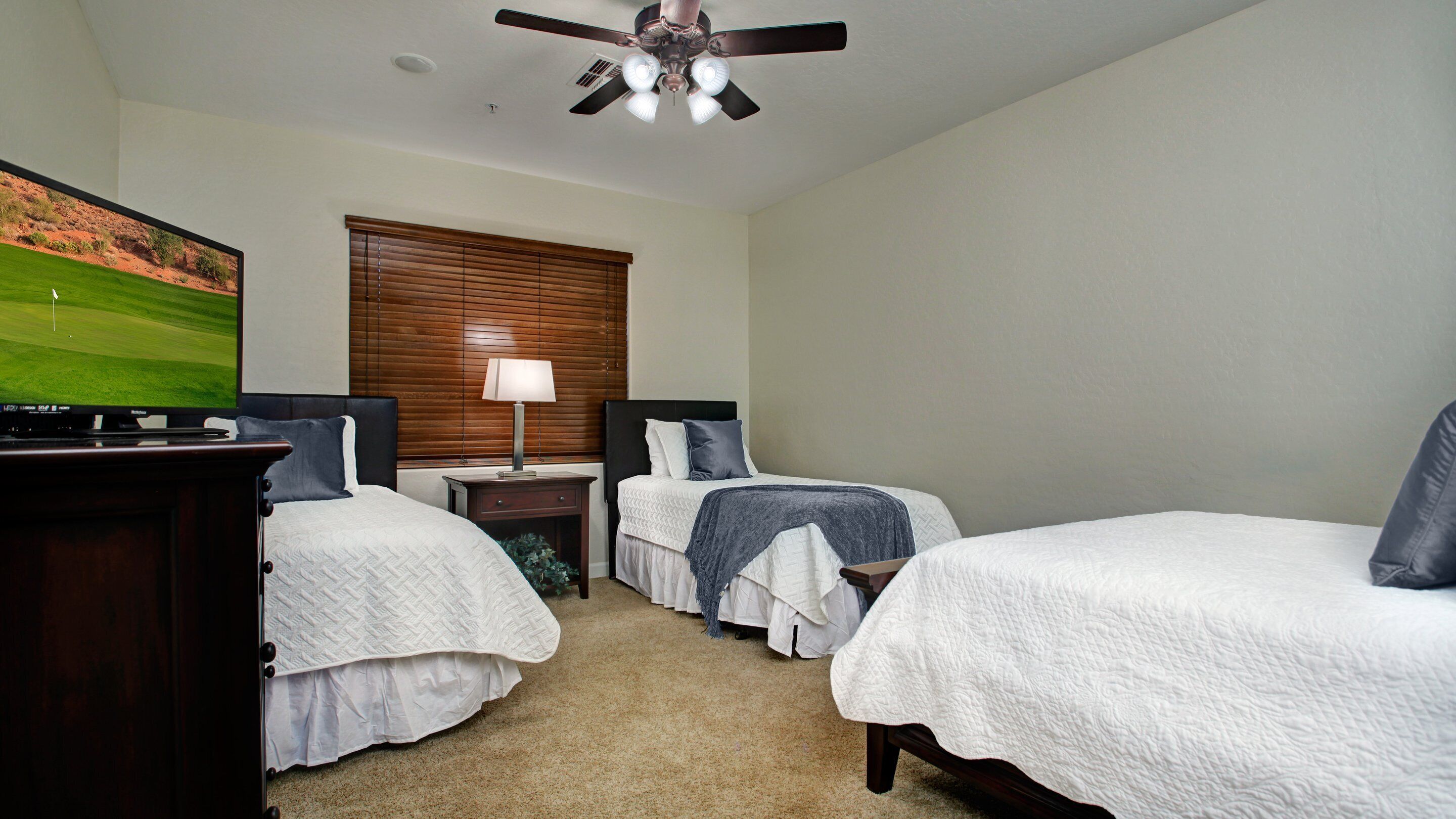 A hotel room with two single beds, a TV, a ceiling fan, and a window with blinds.