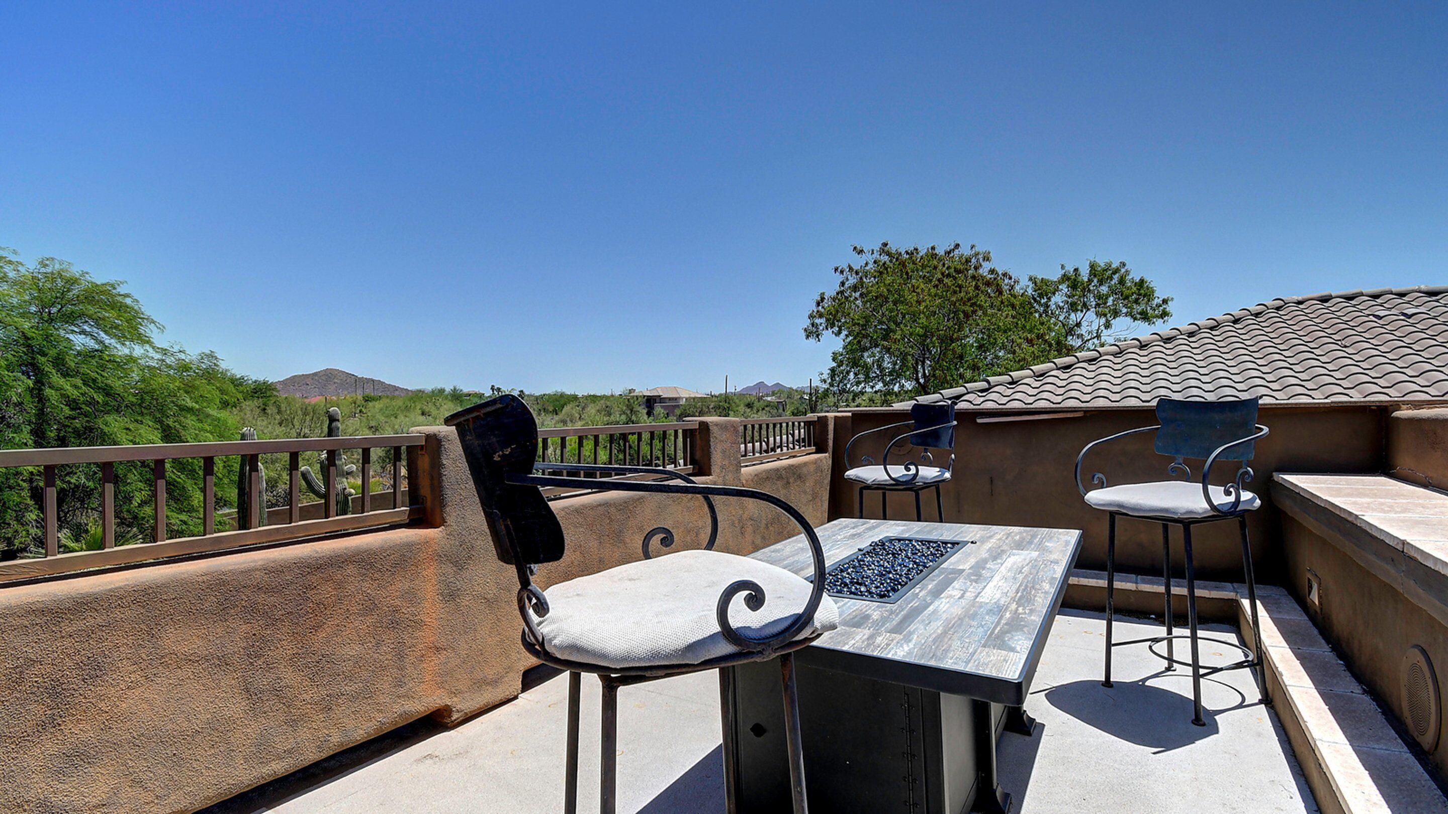 An outdoor patio with a table and two chairs, a railing, and a view of a desert landscape with cacti and distant mountains.