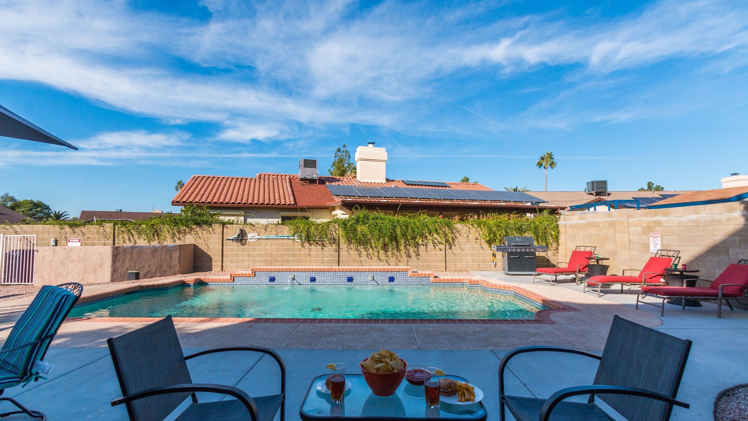 A swimming pool with a tiled deck, surrounded by red lounge chairs and a table with snacks and drinks.