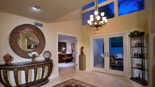 A well-lit hallway with a round mirror, a chandelier, a console table with decorative items, a glass door leading to a dining area, and a metal display cabinet. - The Palm (Scottsdale)