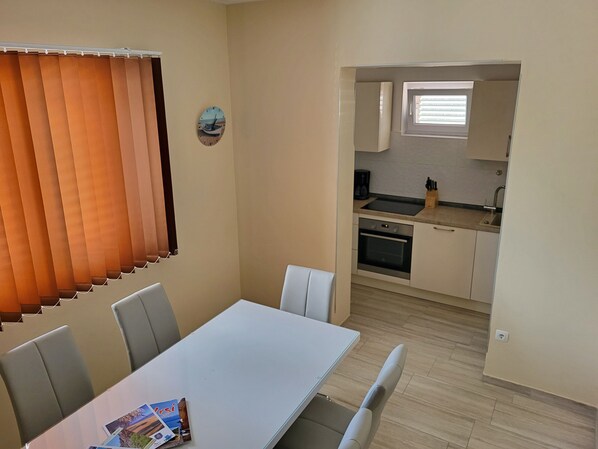 A dining area with a table and chairs, a kitchen with cabinets and appliances, and a window with blinds.