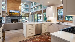 A modern kitchen with a stone fireplace, large windows, and a dining area with a high ceiling. - Impeccable Mountain-Modern Home | Custom-Built Beds | Tesla Charger in Garage (Truckee)
