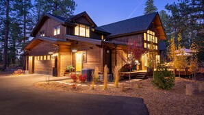 A two-story wooden house with a covered porch, a driveway, and a landscaped yard with trees and shrubs. - Impeccable Mountain-Modern Home | Custom-Built Beds | Tesla Charger in Garage (Truckee)
