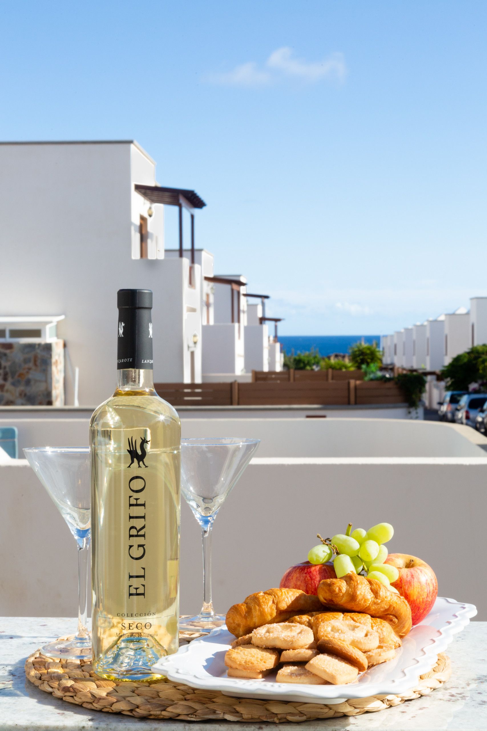 Une bouteille de vin El Grifo, un verre et une assiette de pâtisseries et de fruits, le tout sur une terrasse offrant une vue sur les bâtiments et la mer.
