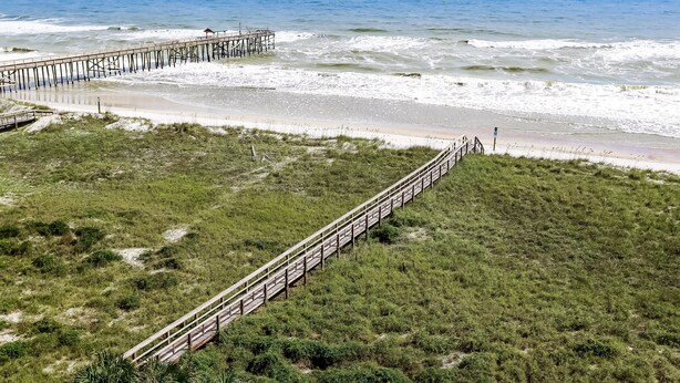 A beach with a wooden pier extending into the ocean, a grassy area with a wooden walkway, and a clear view of the sea. - Ocean and Amelia Island View Condo with Oceanside Pool Access by RedAwning (Fernandina Beach)