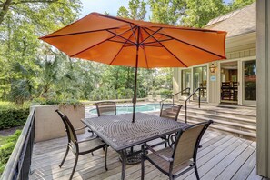 Una terraza de madera con una mesa, sillas y una sombrilla naranja, con una piscina al fondo.