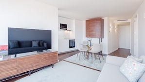 A modern living room with a black sofa, a glass coffee table, and a white couch. There is a dining area with a white table and chairs, and a kitchen with white cabinets and a microwave.