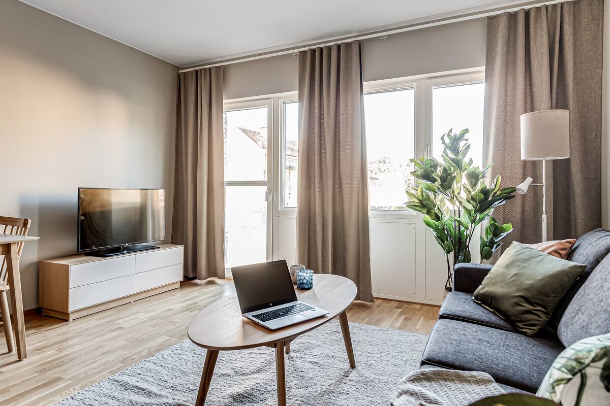 A modern living room with a flat-screen TV, a laptop on a coffee table, a sofa, a lamp, and a large plant.