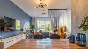 Ein modernes Wohnzimmer mit Esstisch aus Holz, einer gelb gekissenen Couch, einem an der Wand montierten Fernseher und einem großen Fenster mit Vorhängen.