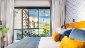 Ein modernes Schlafzimmer mit einem großen Bett, gelb-blauen Kissen, Blick auf eine Stadtlandschaft durch raumhohe Fenster und einem Balkon mit Stuhl und Pflanzen.
