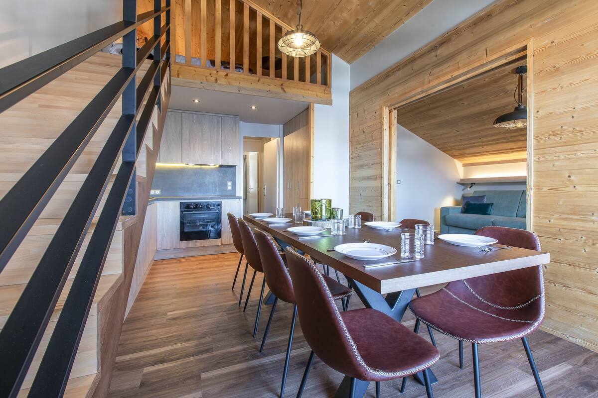Un comedor moderno con una escalera de madera, una cocina al fondo y una zona de estar acogedora.