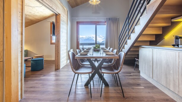 A dining area with a wooden table and chairs, a view of the mountains through a large window, and a staircase with wooden railings. - Vacancéole - Résidence La Duit (Esserts-Blay)