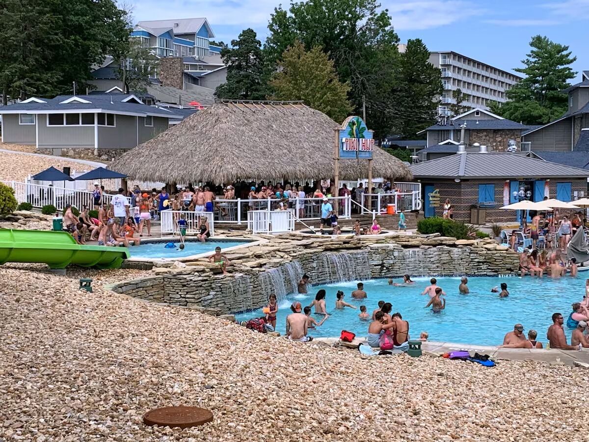 Un espace piscine agrémenté d’une structure à toit de chaume, d’un panneau indiquant « Tiki Hut », et d’une cascade artificielle.