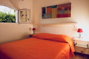 A bedroom with an orange bedspread, a window with a view of greenery, a colorful abstract painting, and two bedside lamps. - Air-conditioned beach house: 7-minute walk, fenced-in yard, charging station for electric cars (Hyères)