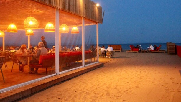 An outdoor dining area with tables and chairs, a covered seating area with red cushions, and a view of the sea.