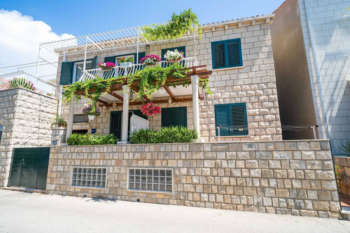 A two-story building with a stone facade, a balcony with hanging plants, and a window with shutters.