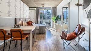 Habitación de hotel moderna con zona de comedor, sofá, televisión y balcón con vistas a la ciudad.