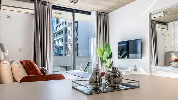 Una habitación de hotel moderna con un ventanal grande, un televisor de pantalla plana, una mesa de comedor con objetos decorativos y un sofá con cojines.