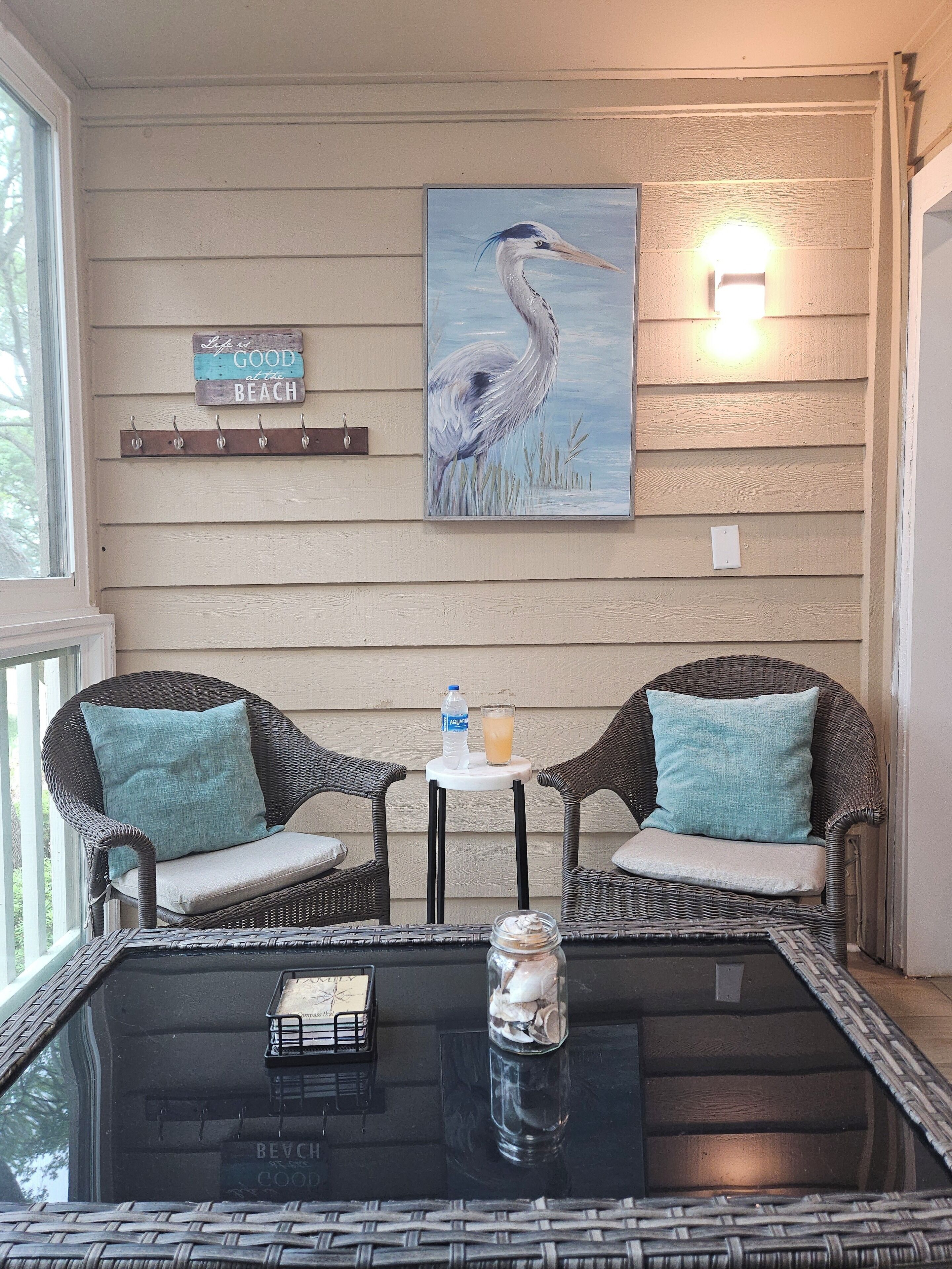 Une pièce avec des chaises en osier, une table en verre, un tableau représentant un oiseau et une pancarte sur laquelle on peut lire : « La vie est belle à la plage ».