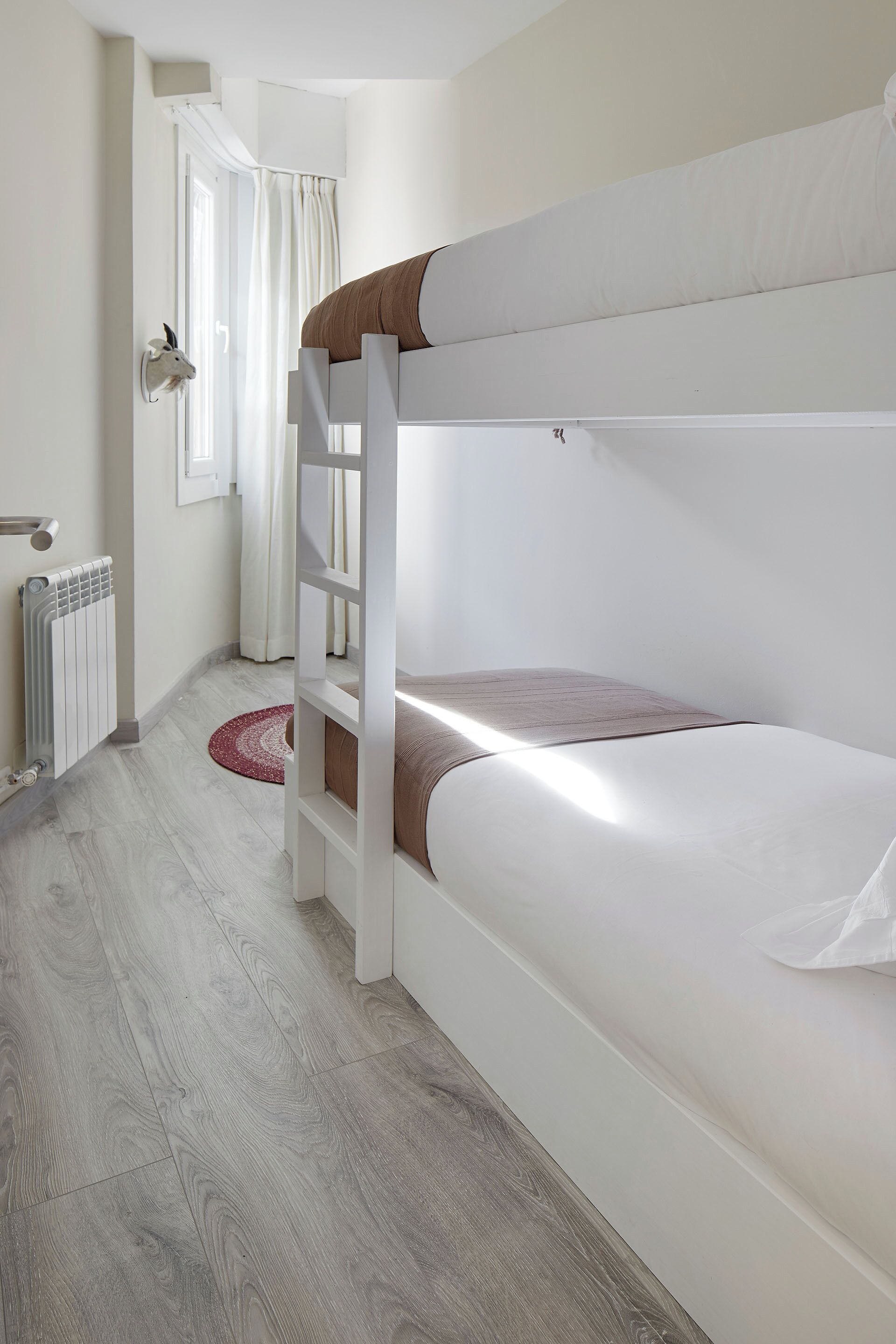 Habitación con literas, piso de madera, radiador, puerta blanca y ventana con cortinas.