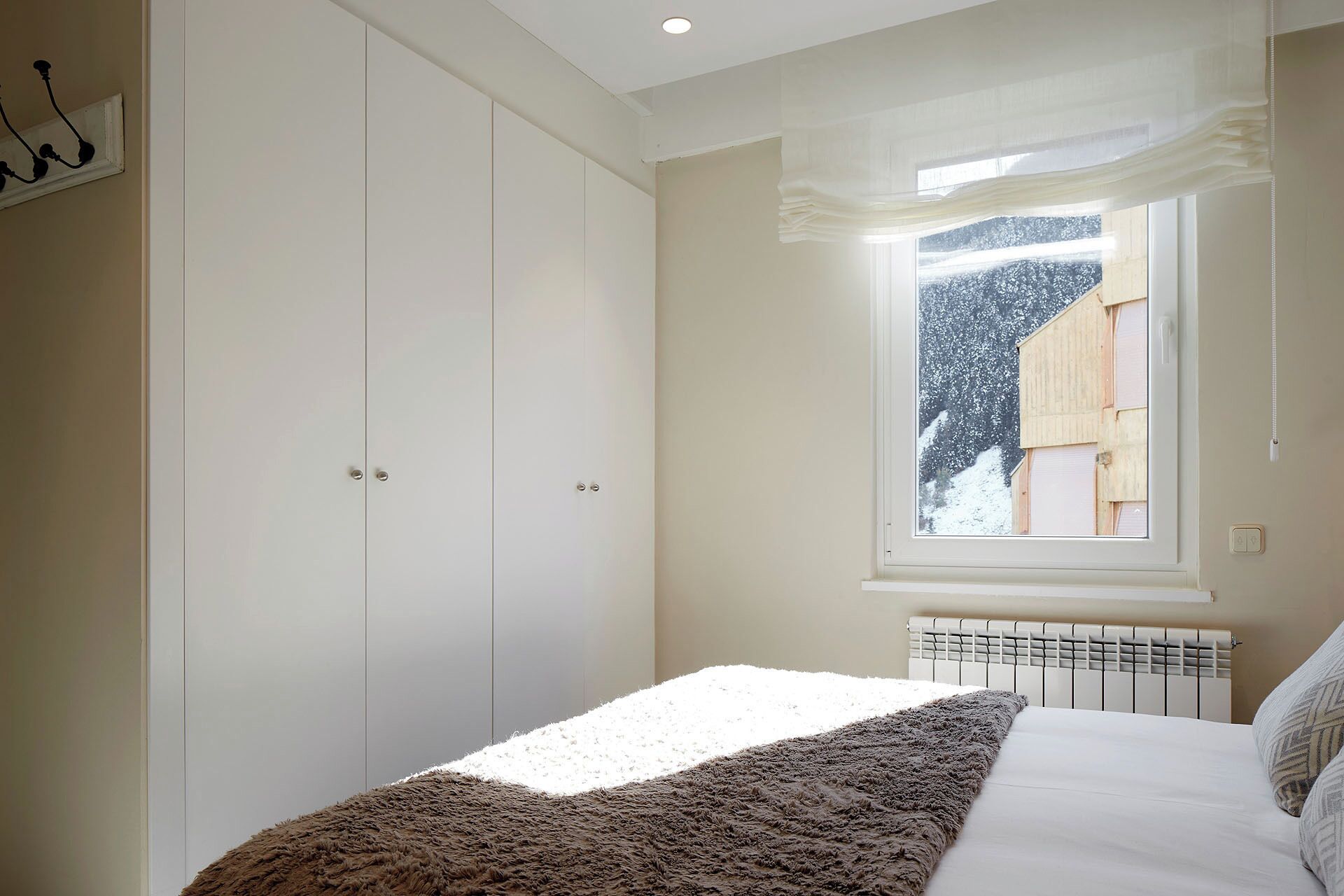 A bedroom with a large wardrobe, a window with a view of a snowy landscape, and a radiator under the window.