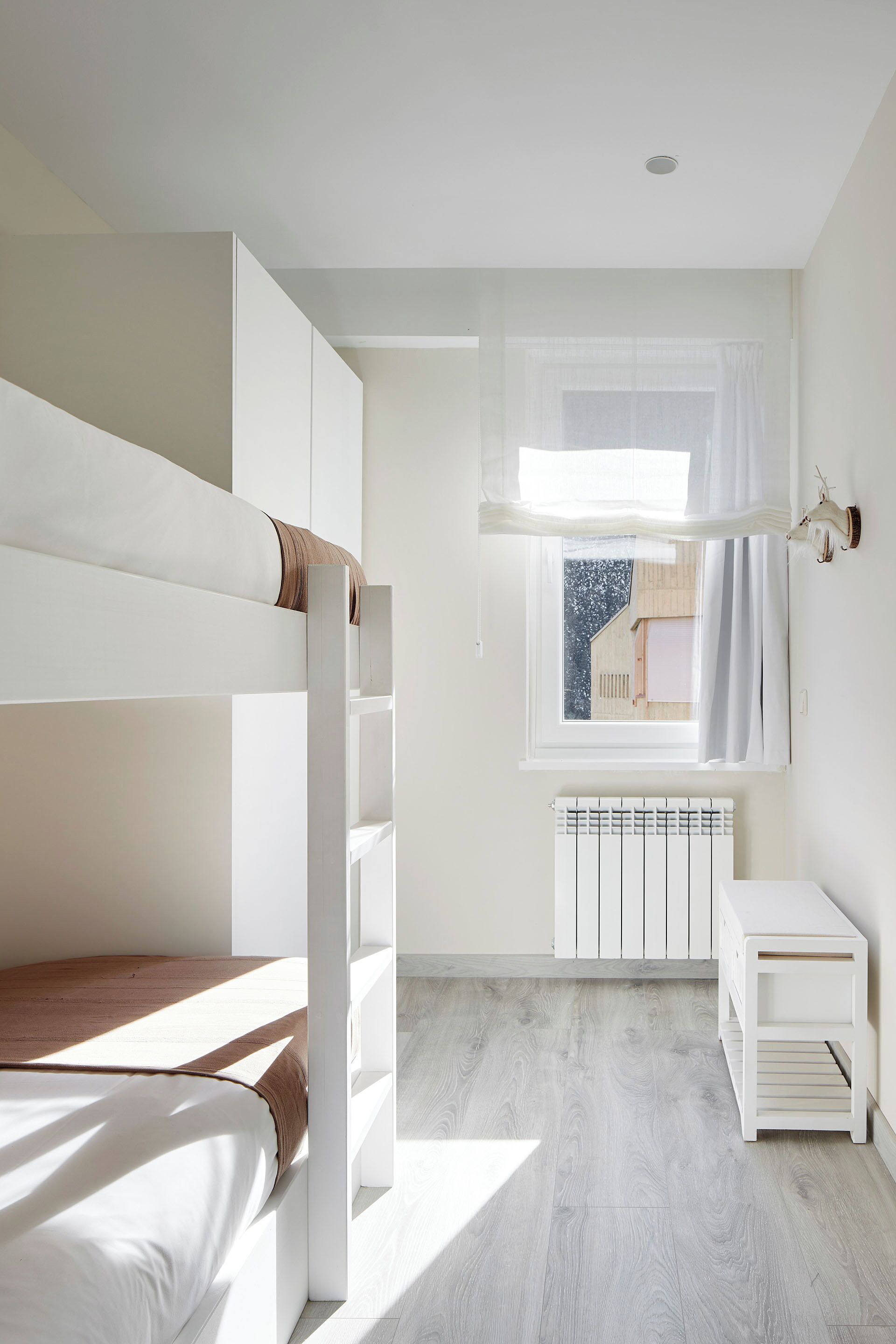 Un lit superposé avec une literie blanche, un radiateur blanc et une petite table de chevet blanche.