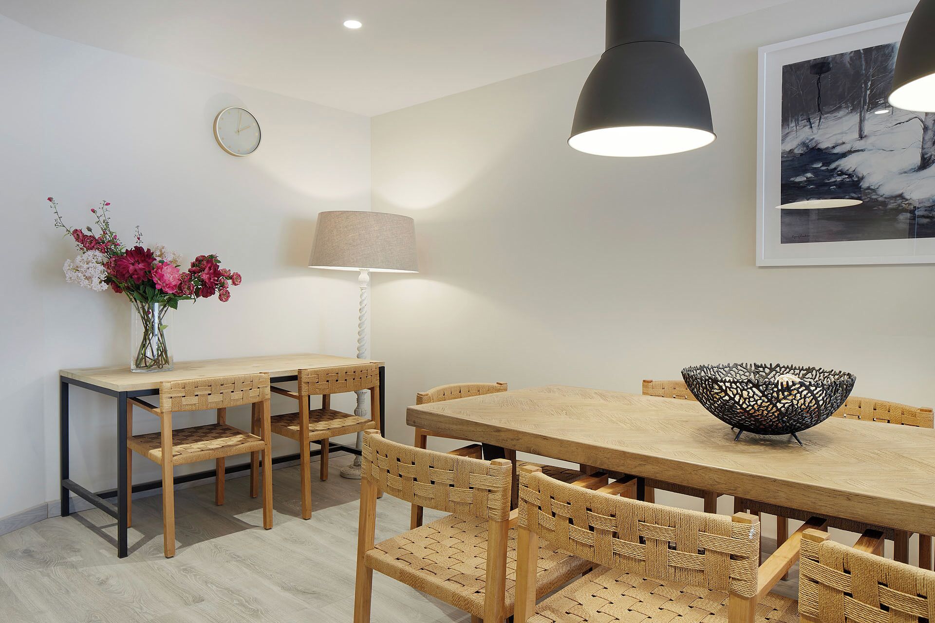 Un comedor con una mesa y sillas de madera, un florero con flores, una lámpara y un cuadro en la pared.