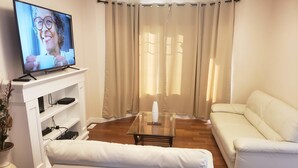 A living room with a white sofa, a glass coffee table, a TV on a stand, and beige curtains. - Stylish Heritage 5 BR House  Ideal For Long Stays - With Jacuzzi (Fort McMurray)