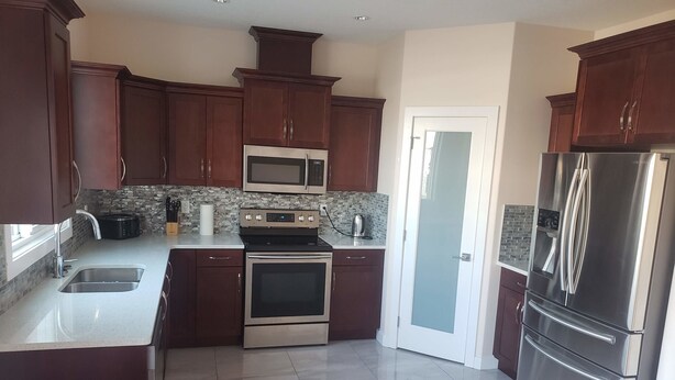 A modern kitchen with dark wooden cabinets, stainless steel appliances, a tiled backsplash, and a glass door leading to another room. - Stylish Heritage 5 BR House  Ideal For Long Stays - With Jacuzzi (Fort McMurray)