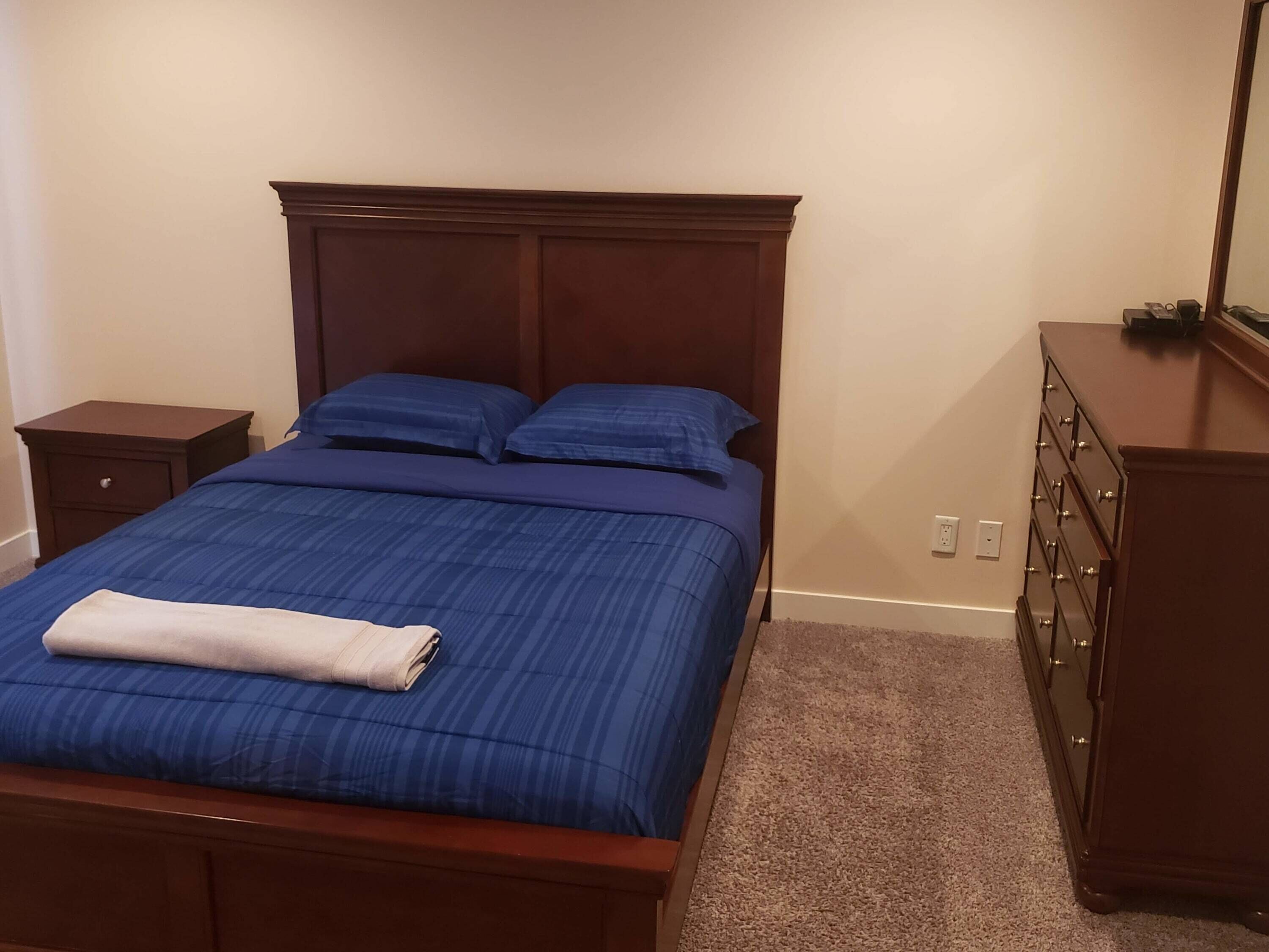 Een slaapkamer met een houten bed, blauw beddengoed, een nachtkastje, een dressoir en een vloerbedekking.