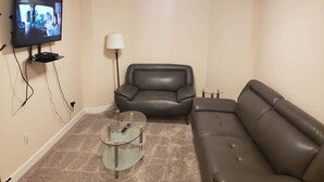 Ein Wohnzimmer mit an der Wand montiertem Fernseher, einem Ledersofa, einem Glastisch und einer Lampe.