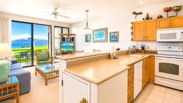 A modern kitchen with a large island, wooden cabinets, and a view of the sea through the sliding glass doors.