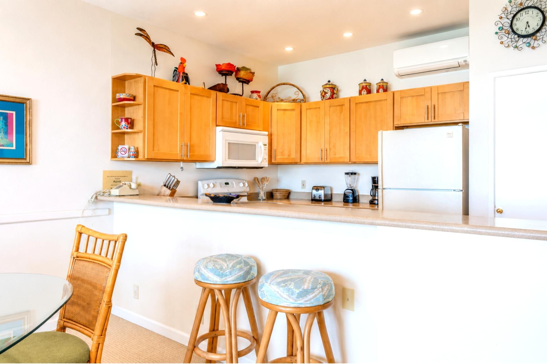 A kitchen with wooden cabinets, a microwave, a toaster, a blender, and a refrigerator. There are two bar stools with patterned seats.