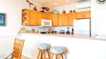 A kitchen with wooden cabinets, a microwave, a toaster, a blender, and a refrigerator. There are two bar stools with patterned seats.