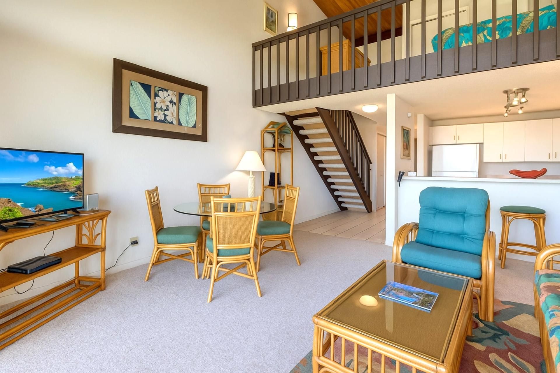 A living room with a TV, a dining table with chairs, a sofa, a staircase, and a kitchen area.