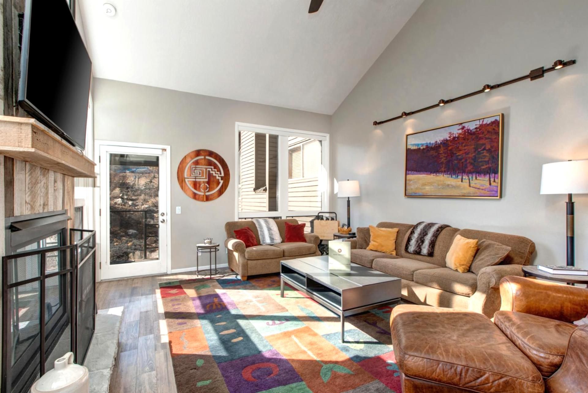A spacious living room with a large sofa, a coffee table, and a colorful rug. There is a television on a wooden stand, a painting on the wall, and a window with blinds.