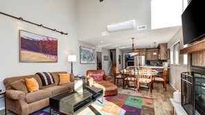 A modern living room with a sofa, coffee table, and colorful rug. There is a dining area with a table and chairs, and a kitchen with cabinets and appliances.