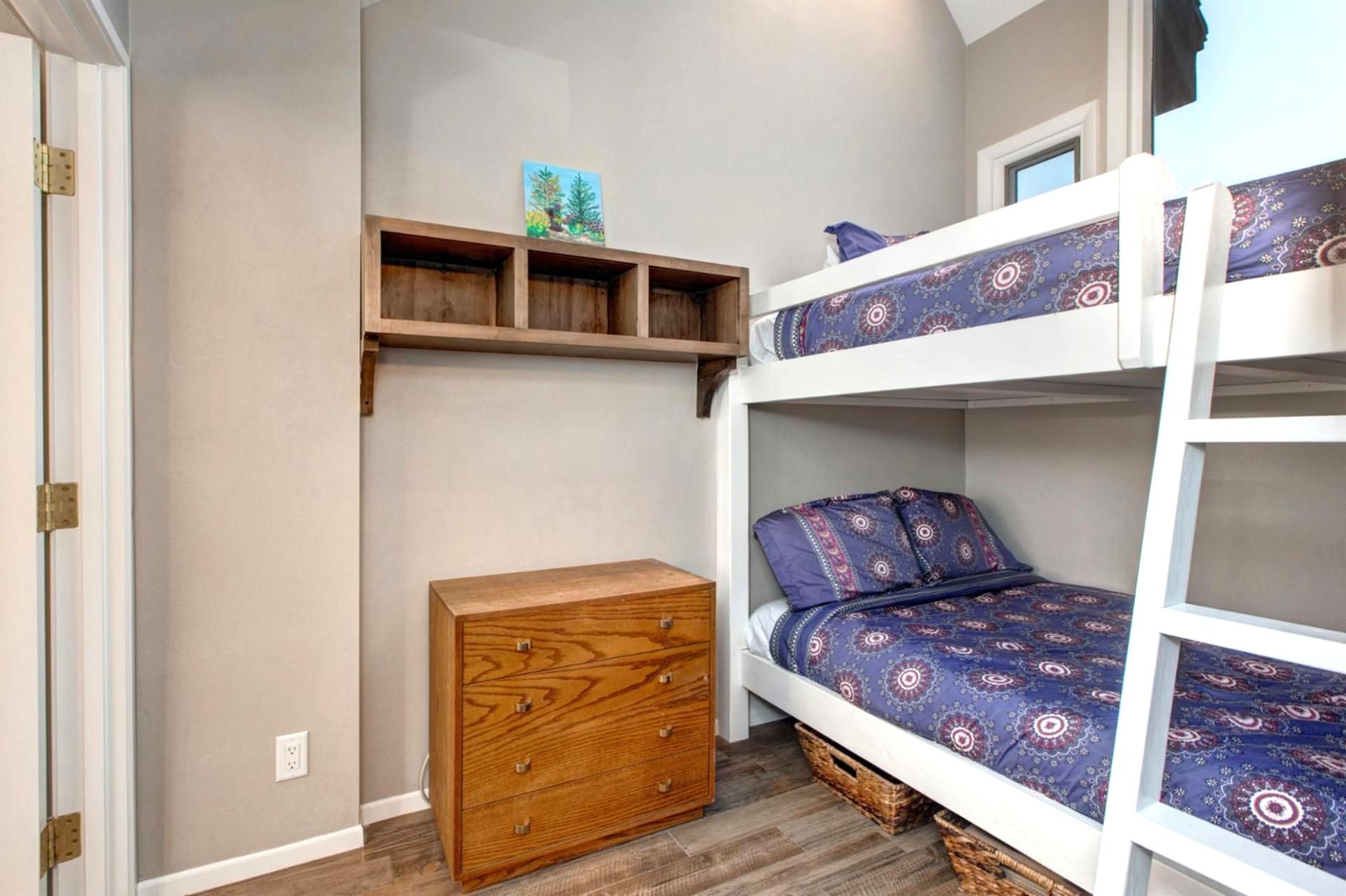 A bunk bed with patterned bedding, a wooden dresser, and a shelf with a small painting.